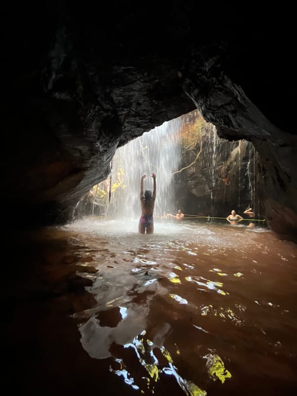 Silhueta de uma pessoa de braços erguidos sob a queda, vista de dentro da gruta escura da Caverna