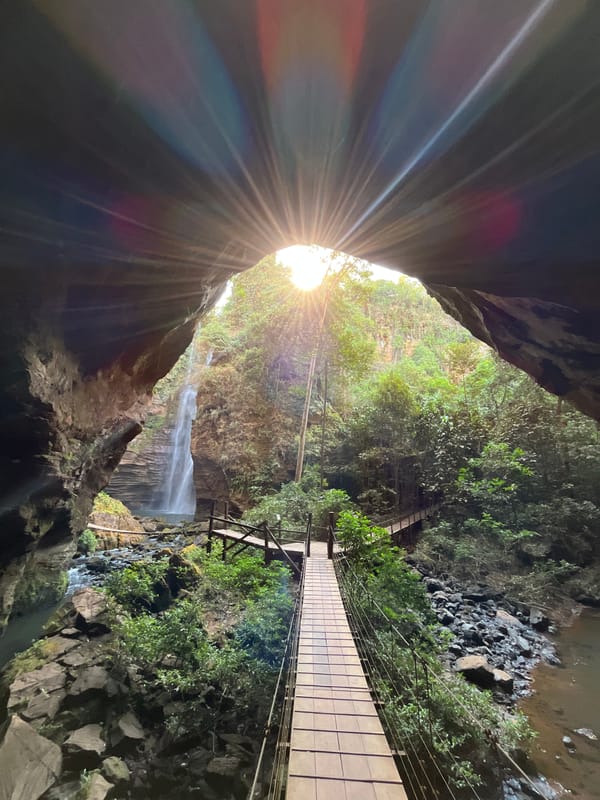Passarela de madeira levando à gruta, com o sol estourando na abertura do alto e uma queda fina ao lado