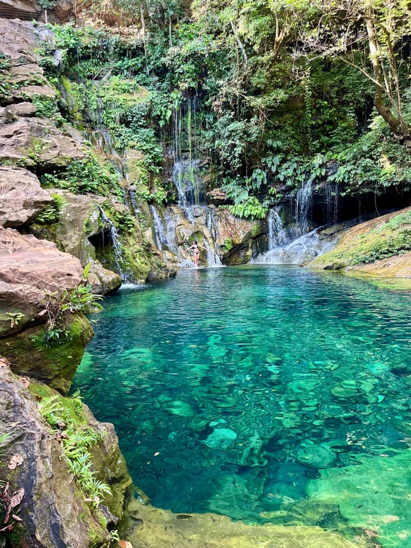 Poço verde-turquesa transparente com quedas finas descendo pela parede de pedra e pela mata