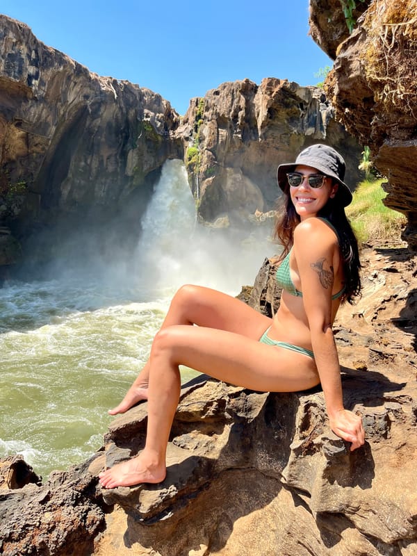 Mulher de chapéu sentada numa pedra, sorrindo, com a Cachoeira do Prata atrás