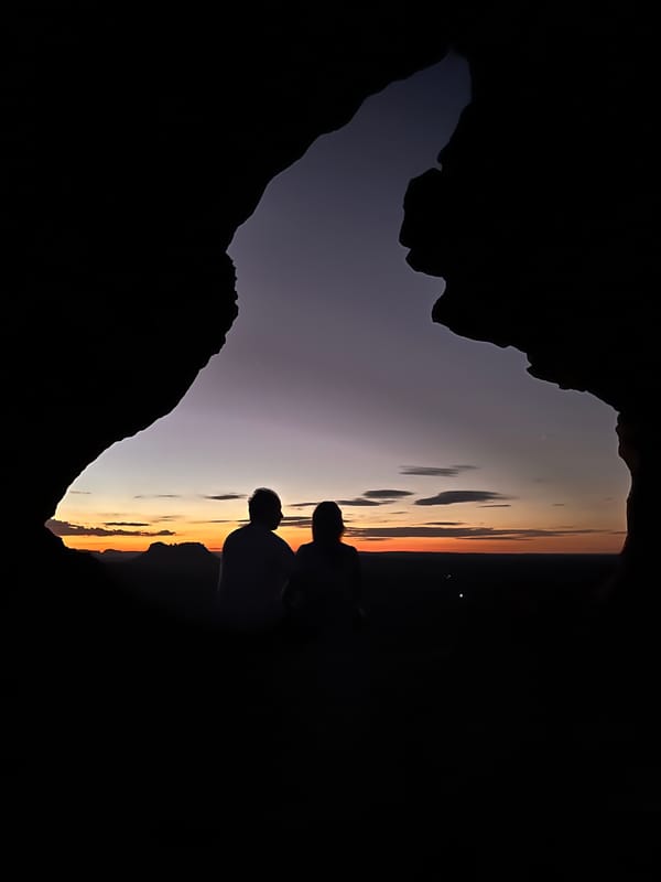 Silhueta de duas pessoas no buraco do Portal, com o céu do fim de tarde recortado pela pedra