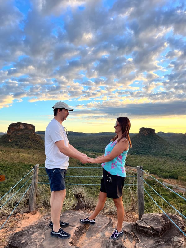 Casal de mãos dadas no mirante do Portal da Chapada, sob um céu de nuvens, com as mesas ao fundo