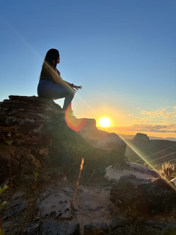 Silhueta de uma mulher sentada no alto de uma pedra, com o sol baixo estourando ao lado