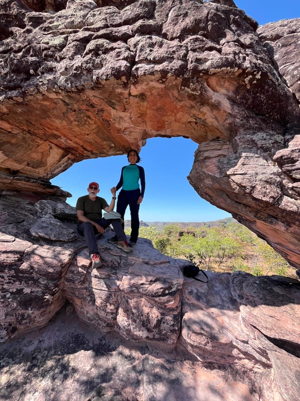 Duas pessoas sob um grande arco de arenito, uma sentada e outra de pé