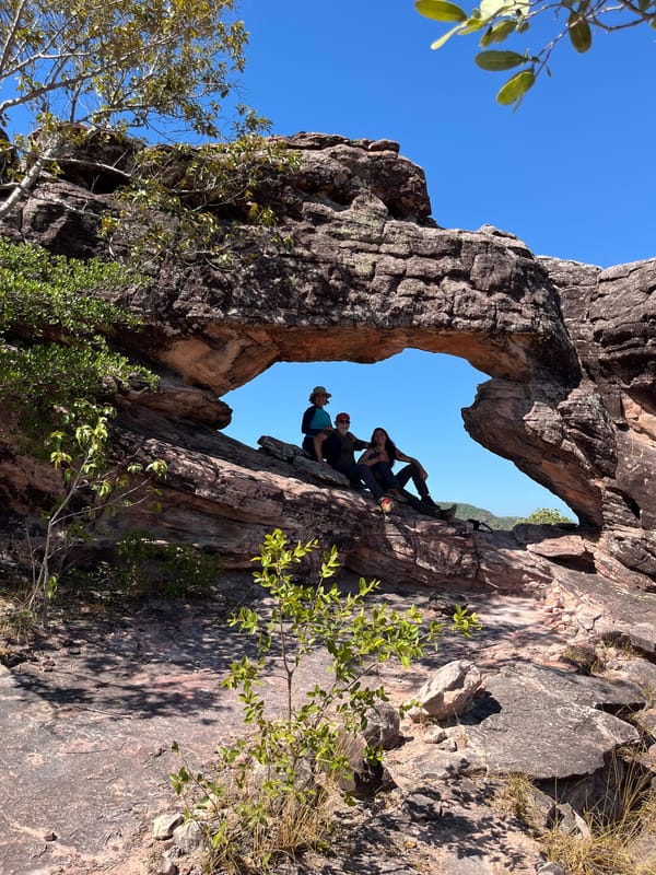 Duas pessoas sentadas sob o vão de um grande arco de arenito, com o céu azul ao fundo
