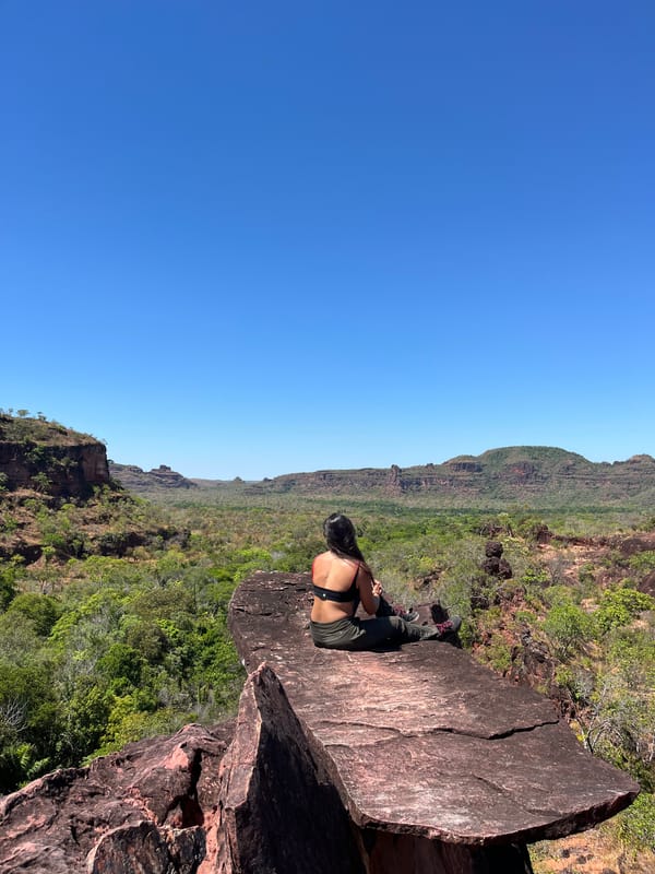 Mulher sentada na ponta de uma laje de pedra, olhando o cerrado e as mesas ao longe