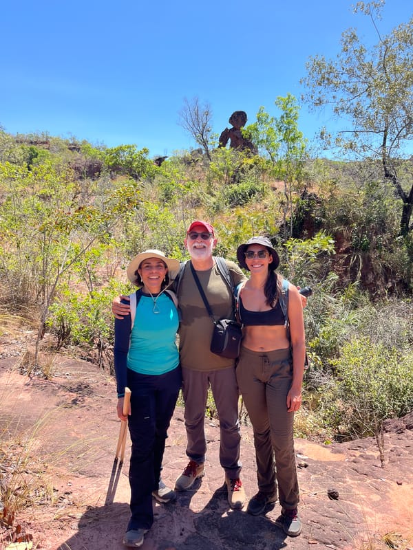 Três caminhantes abraçados na trilha de terra, com uma pedra em forma de cogumelo atrás