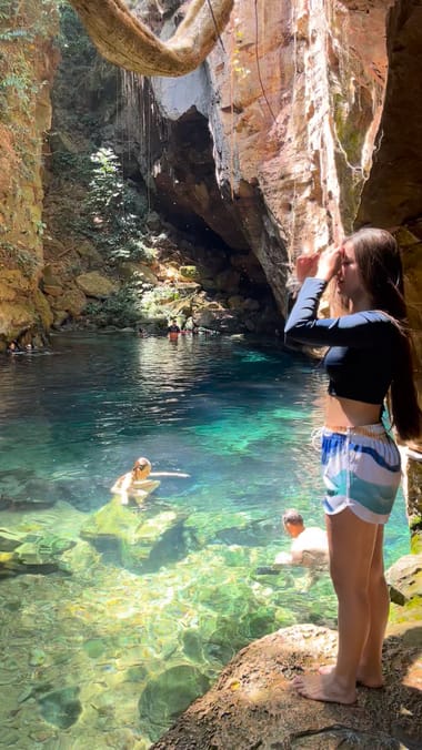 Mulher de pé numa pedra à beira do poço turquesa dentro da gruta, com gente nadando